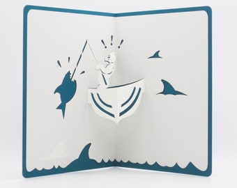 May include: A white pop-up card with a blue border. The card features a 3D design of a person fishing in a boat with a shark jumping out of the water. The shark is blue and the boat is white with blue stripes. There are three blue shark fins in the background.