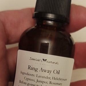 Pode incluir: Uma garrafa de vidro castanho com um conta-gotas preto e um rótulo branco que diz "Jamilah's Naturals Ring Away Oil". O rótulo lista os ingredientes: lavanda, helichrysum, cipreste, zimbro e alecrim. O rótulo também inclui instruções para usar o óleo: "Antes de ir para a cama, coloque 6 gotas em uma bola de algodão. Coloque a bola de algodão no seu ouvido durante a noite. Se a bola de algodão for desconfortável, você pode aplicar o óleo no lóbulo da sua orelha, atrás da sua orelha ou na parte de trás do seu pescoço. Isso não é destinado a diagnosticar, tratar, curar ou prevenir qualquer doença."