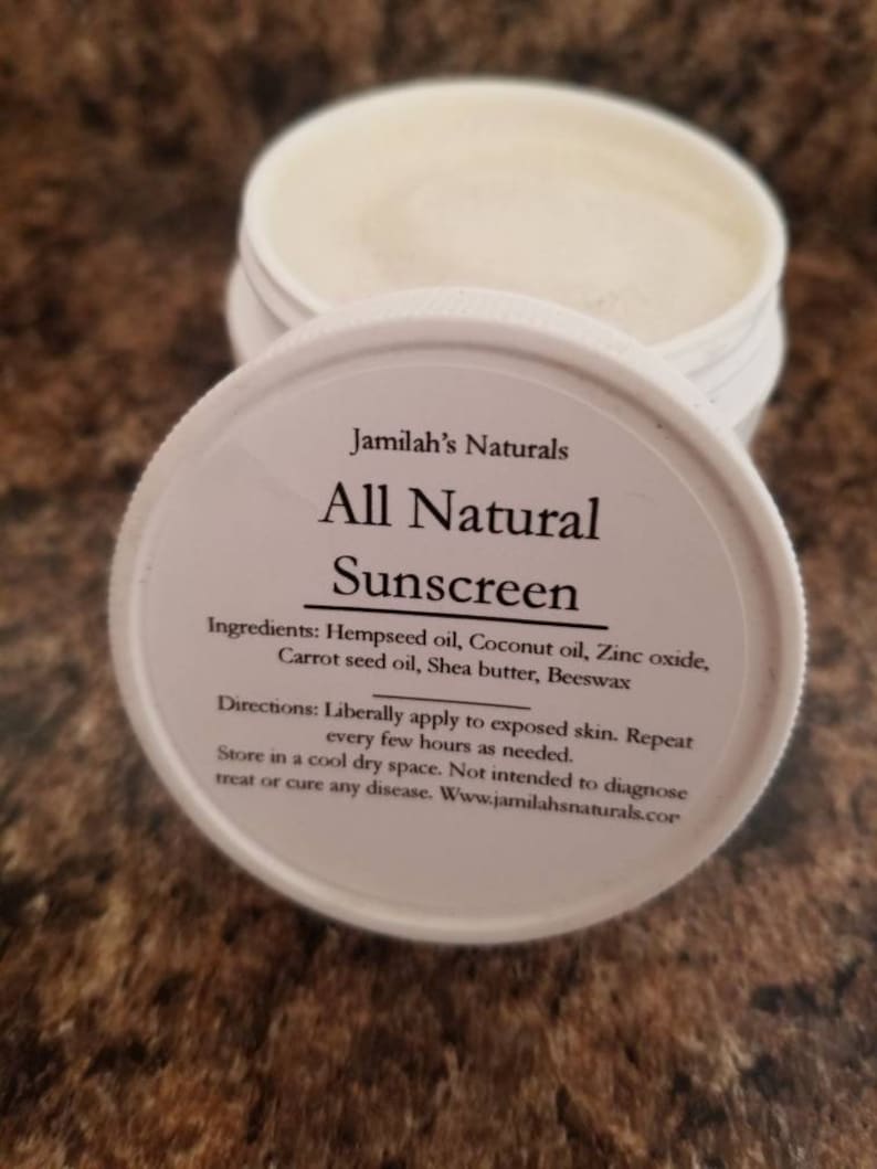 May include: A white jar of Jamilah's Naturals All Natural Sunscreen with the lid open. The lid displays the product name and ingredients: Hempseed oil, Coconut oil, Zinc oxide, Carrot seed oil, Shea butter, Beeswax.