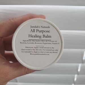 May include: White jar with a black label that reads "Jamilah's Naturals All Purpose Healing Balm". The label lists ingredients including shea butter, olive oil, coconut oil, beeswax, lavender, rosemary, peppermint, and vitamin E. The label also includes directions for use and a website address.