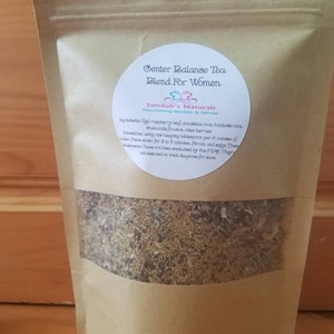 May include: A brown paper bag with a white label that reads "Center Balance Tea Blend For Women" and "Jamilah's Naturals Nourishing Bodies & Minds". The label also lists the ingredients and directions for use. The bag contains a blend of dried herbs.