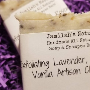 May include: Close-up of two handmade soap bars with white labels. The labels read "Jamilah's Naturals" and "Exfoliating Lavender, Vanilla Artisan Cl". The soaps are light in color and speckled with dark flecks, resting on purple shredded paper.