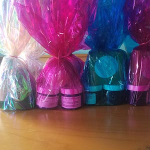 May include: Four sets of small jars wrapped in colorful cellophane. Each set contains two jars, one purple and one blue. The jars are wrapped in iridescent cellophane in shades of pink, blue, and white.