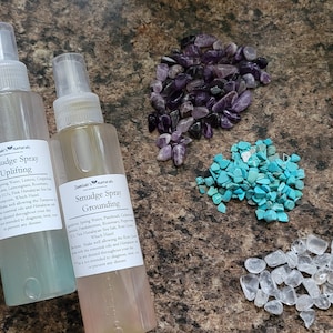 Könnte beinhalten: Drei durchsichtige Glasflaschen mit Sprühköpfen, die mit "Smudge Spray" und den Düften "Klarheit", "Aufbahnung" und "Erdung" beschriftet sind. Die Flaschen sind auf einer braunen, strukturierten Oberfläche mit kleinen Haufen Amethyst, Türkis, rosa Himalaya-Salz und klarem Quarz angeordnet.
