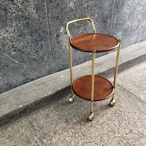 Op de afbeelding: Een verrijdbare trolley met twee niveaus en twee ronde houten planken in een warme bruine afwerking. Het frame is gemaakt van goudkleurige metalen buizen, inclusief een gebogen handvat aan de bovenkant en drie kleine wieltjes aan de onderkant.