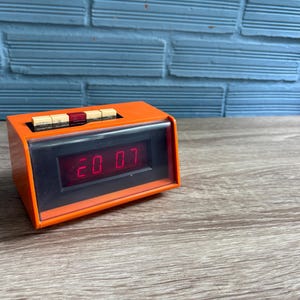 Puede incluir: Un despertador digital naranja que muestra la hora "20 07" en números rojos. El reloj tiene forma rectangular con una pantalla negra y botones en la parte superior. Está sobre una superficie de madera.