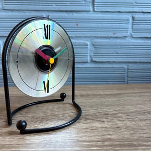 Puede incluir: Un reloj de escritorio único hecho de un CD reutilizado. La esfera del reloj presenta números romanos y manecillas coloridas. El reloj está sostenido por un soporte de metal negro, agregando un toque moderno al diseño. Se encuentra sobre una superficie de madera.