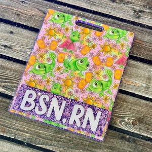 May include: A rectangular badge holder with a purple glitter border and a hole at the top. The design features a pink background with green chameleons, yellow suns, and orange lanterns. The text "BSN RN" is displayed in white.
