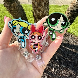 May include: Three glittery acrylic badge reels featuring the Powerpuff Girls characters Bubbles, Blossom, and Buttercup. Each character is depicted in their signature color scheme of blue, pink, and green with a clear plastic retractable cord.