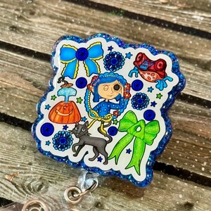 May include: A badge reel with a blue glitter border, showcasing a colourful illustration. The design includes a girl, a black cat, bows, and other whimsical elements in blue, green, orange, and red. The reel has a clear plastic base and a metal clip.