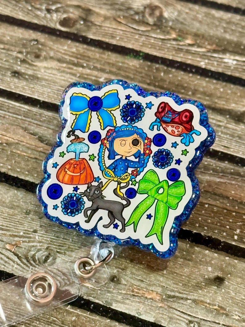 May include: A badge reel with a blue glitter border, showcasing a colourful illustration. The design includes a girl, a black cat, bows, and other whimsical elements in blue, green, orange, and red. The reel has a clear plastic base and a metal clip.