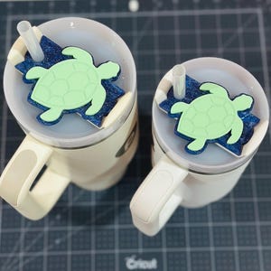 May include: Two white travel mugs with handles, each topped with a lid and a straw. The lids feature a blue glittery base and a light green turtle-shaped charm. The mugs are on a dark surface.