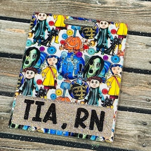 Peut inclure: Un porte-badge rectangulaire avec une bordure scintillante et le texte "TIA. RN". Le badge pr&eacute;sente un motif color&eacute; de personnages de dessins anim&eacute;s et de boutons. Le fond est une surface en bois.