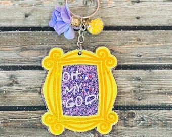 May include: A yellow glittery keychain featuring a frame design with the text "OH. MY. GOD!". The keychain is adorned with a purple flower, a gold bead, and a yellow pom-pom. The background is a wooden surface.