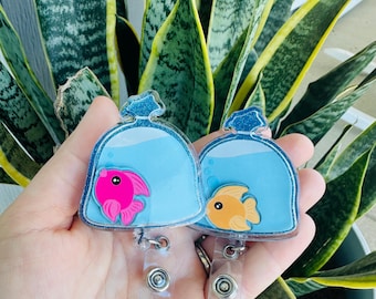 May include: Two fishbowl-shaped badge reels. One reel features a pink fish, the other an orange fish, both inside a blue fishbowl design. The reels have a clear plastic strap and a silver metal clip.