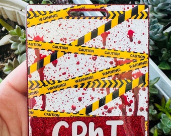 Puede incluir: Un porta-credenciales rectangular con un borde rojo brillante. El diseño presenta cinta de precaución amarilla y negra, salpicaduras rojas y el texto "WARNING!" y "CAUTION!". La sección inferior tiene las letras "CPhT" en blanco.