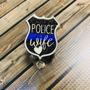 May include: A badge-shaped retractable ID holder with the words "POLICE wife" in white script over a blue stripe. The badge has a black glitter background and a white heart. The holder is attached to a clear plastic clip.