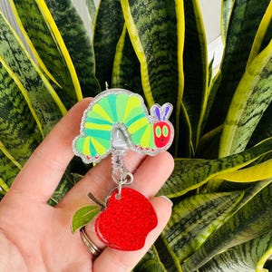 May include: A colorful badge reel featuring a green and yellow caterpillar design with glitter accents. The reel includes a red glitter apple charm with a green leaf. The background is a plant with green and yellow leaves.