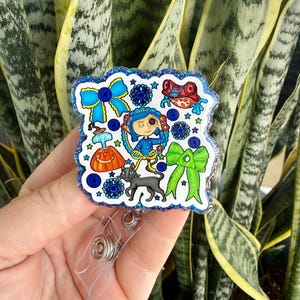 May include: A badge reel with a cartoon illustration. The design includes a blue bow, a girl, a black cat, and other whimsical elements. The badge reel has a clear plastic clip and a retractable cord. The design is outlined with glitter.