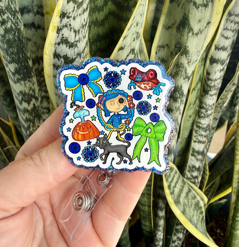 May include: A badge reel with a cartoon illustration. The design includes a blue bow, a girl, a black cat, and other whimsical elements. The badge reel has a clear plastic clip and a retractable cord. The design is outlined with glitter.
