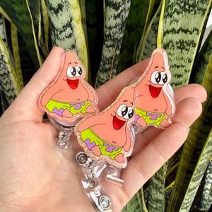 May include: Three acrylic badge reels featuring a glittery finish and a cartoon character design of a pink starfish wearing lime green shorts with purple flower patterns. Each badge reel has a clear plastic strap and a metal snap button closure.