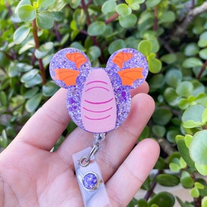 May include: A purple glitter badge reel featuring a cartoon dragon design with orange wings and a pink face. The badge reel has a clear plastic strap and a purple glitter button.