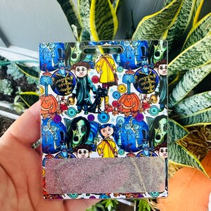 Peut inclure: Un porte-badge rectangulaire avec un motif color&eacute; repr&eacute;sentant des personnages et des images d'un film fantastique. Le motif comprend des citrouilles, des boutons et diverses figures sur fond bleu. La partie inf&eacute;rieure pr&eacute;sente un accent rectangulaire scintillant.
