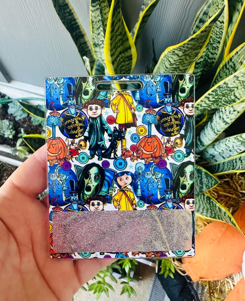 Peut inclure: Un porte-badge rectangulaire avec un motif color&eacute; repr&eacute;sentant des personnages et des images d'un film fantastique. Le motif comprend des citrouilles, des boutons et diverses figures sur fond bleu. La partie inf&eacute;rieure pr&eacute;sente un accent rectangulaire scintillant.
