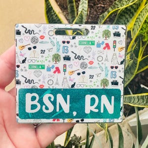 May include: A rectangular badge holder with a teal glittery bottom section displaying the text "BSN, RN" in white. The top section has a white background with various small illustrations, including hearts, cats, and the Eiffel Tower.