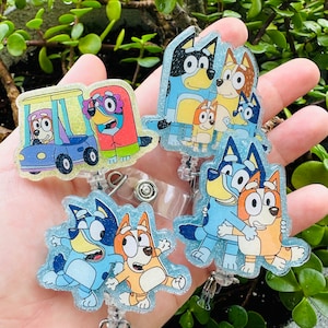 May include: Several glittery badge reels featuring cartoon characters in various poses. The characters are in shades of blue, orange, and red. Each reel has a clear plastic clip and a retractable cord.