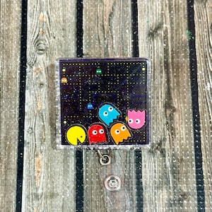 May include: A square, glittery badge reel featuring a Pac-Man-themed design. The design includes Pac-Man and the ghosts on a black background with a maze pattern. The reel has a clear plastic clip and a silver border.