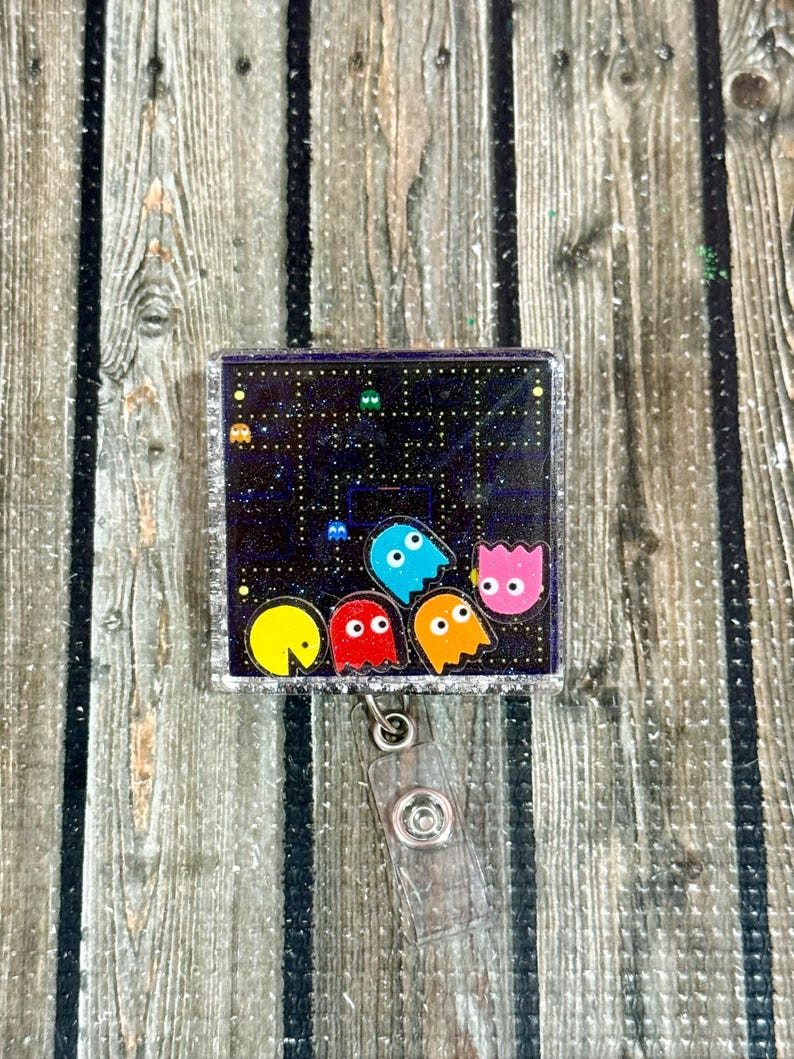 May include: A square, glittery badge reel featuring a Pac-Man-themed design. The design includes Pac-Man and the ghosts on a black background with a maze pattern. The reel has a clear plastic clip and a silver border.