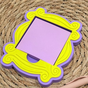 May include: A Friends-themed memo pad holder in a yellow and purple frame design. The frame holds a square of purple sticky notes. A light pink pen rests on the woven surface next to the memo pad holder.