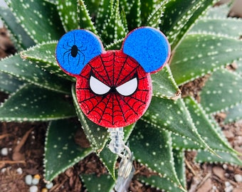 May include: A red, white, and blue Spider-Man themed badge reel. The badge reel is shaped like Mickey Mouse ears, with a spider web design on the face and a small black spider on one ear. The reel is attached to a clear plastic clip.