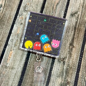 May include: A square, clear plastic retractable badge reel featuring a pixelated Pac-Man design. The design includes yellow, red, blue, orange, and pink ghosts against a black background with white dots. The reel has a silver glitter border and a clear plastic clip.