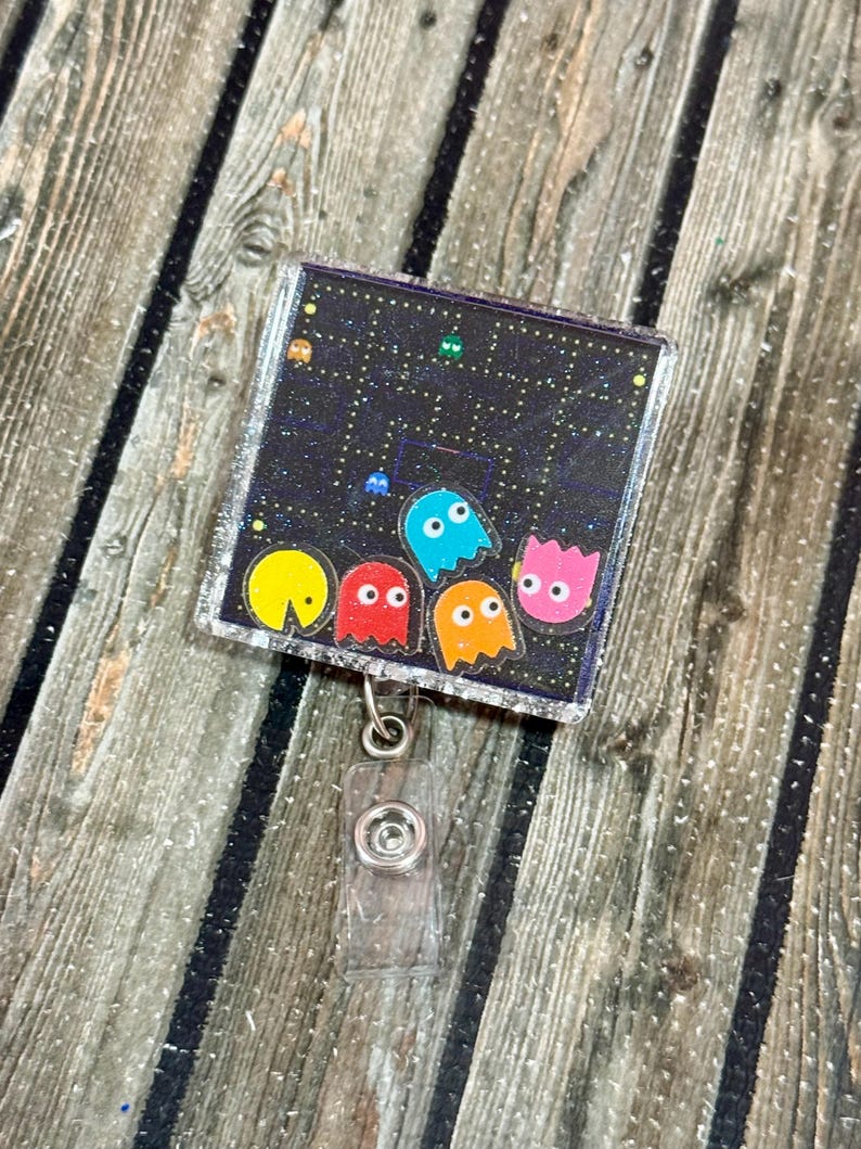 May include: A square, clear plastic retractable badge reel featuring a pixelated Pac-Man design. The design includes yellow, red, blue, orange, and pink ghosts against a black background with white dots. The reel has a silver glitter border and a clear plastic clip.