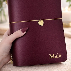 A hand holding a burgundy leather-bound journal with a gold heart charm and the name "Mia" printed on the cover.