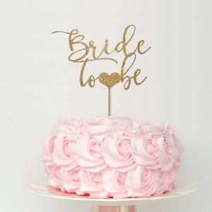 May include: A pink cake with a gold glitter "Bride to Be" cake topper. The cake is decorated with pink frosting in a rosette pattern.