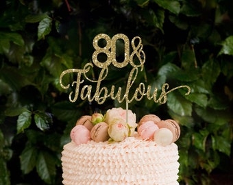 May include: A pink and white cake with a gold glitter topper that says "80 & Fabulous". The cake is decorated with macarons and is on a light blue cake stand. There are also small desserts with fruit and whipped cream on a table in front of the cake.