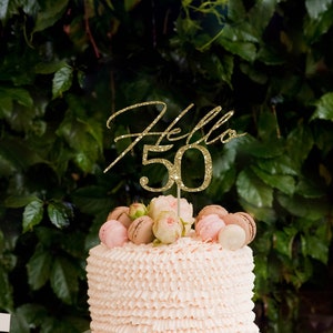 May include: A pink and white cake with a "Hello 50" topper, surrounded by small desserts and flowers on a table.