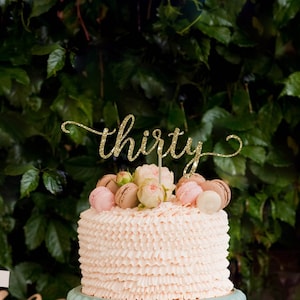 May include: A pink and white cake with a gold glitter topper that says "thirty" on a light blue cake stand. The cake is surrounded by small desserts and flowers.