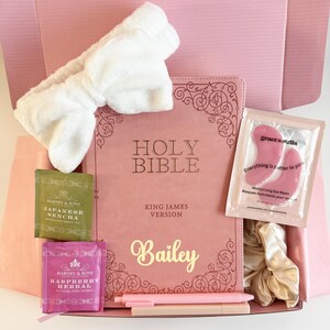 Könnte beinhalten: Eine rosa Geschenkbox mit einer rosa Bibel mit dem Namen "Bailey" in goldener Schrift, einem weißen Stirnband, Teesachets, Augenmasken, Stiften und einem goldenen Haargummi. Die Bibel trägt die Aufschrift "Holy Bible, King James Version."