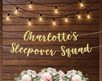 May include: Gold glitter banner with the text "Charlotte's Sleepover Squad" hanging on a wood wall with string lights. The banner is strung with white thread.  There are flowers in the foreground.