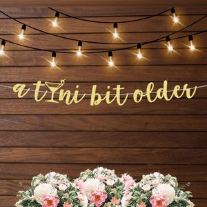 May include: A gold glitter banner with the text "a tini bit older" hanging on a wood wall with string lights. The banner features a gold glitter martini glass.