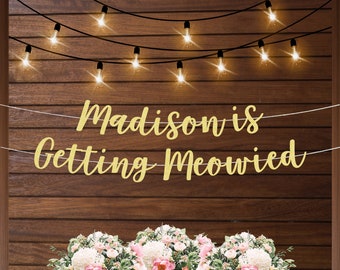 May include: Gold glitter banner with the text "Madison is getting Meowed" hanging on a wood wall with string lights. The banner is in front of a bouquet of white and pink flowers.