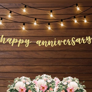 May include: Gold glitter banner with the text "happy anniversary" hanging on a wood wall with string lights.