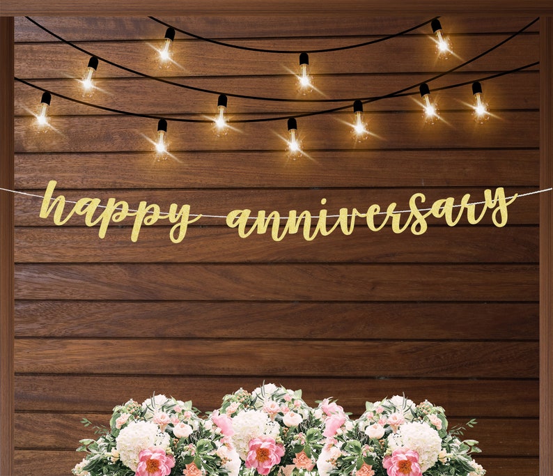 May include: Gold glitter banner with the text "happy anniversary" hanging on a wood wall with string lights.