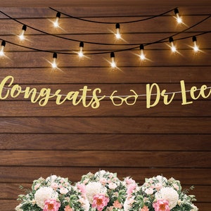 May include: Gold glitter banner with the text "Congrats & Dr Lee" hanging on a wood wall with string lights. The banner is decorated with a pair of gold glitter glasses.