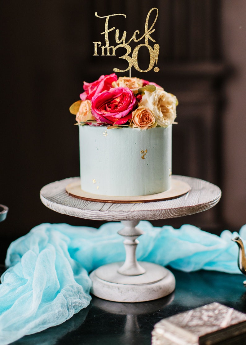 Puede incluir: Un pastel azul claro con motas doradas y rosas rosadas y crema en la parte superior. Un adorno de pastel de purpurina dorada dice "Fuck I'm 30!" El pastel est&aacute; en un soporte de pastel blanco.