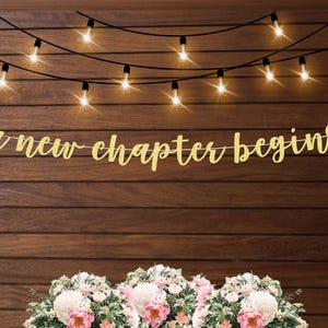 May include: A gold glitter banner with the text "a new chapter begins" hanging on a wood wall with string lights.